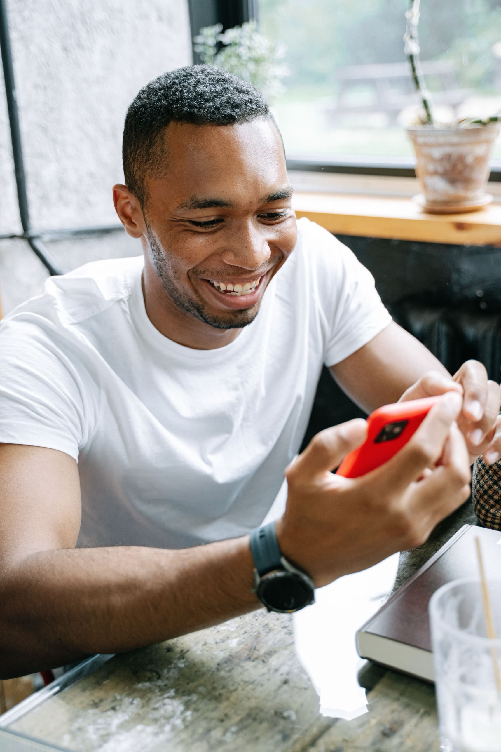 A man laughing at his phone across a cafe table from someone, by a window.