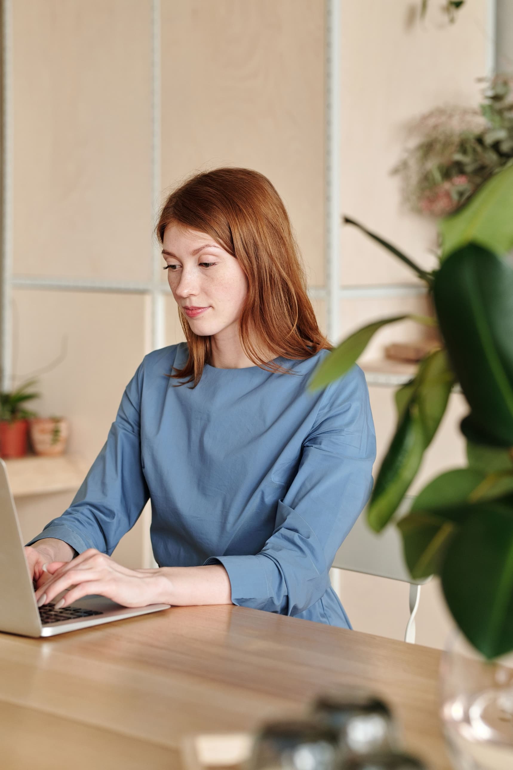 A woman in a blue dress working at a laptop in a calm room, plants around her.
