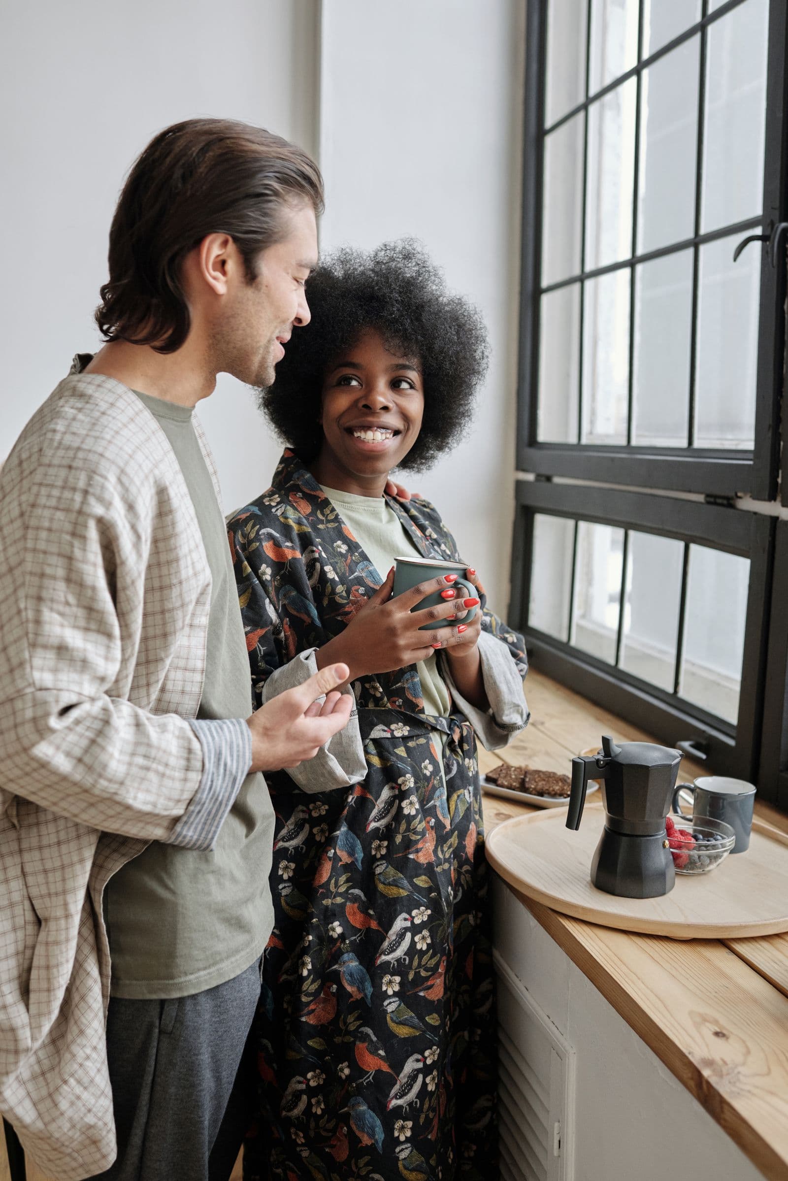 Two people standing together at a kitchen window in the morning, laughing over coffee.
