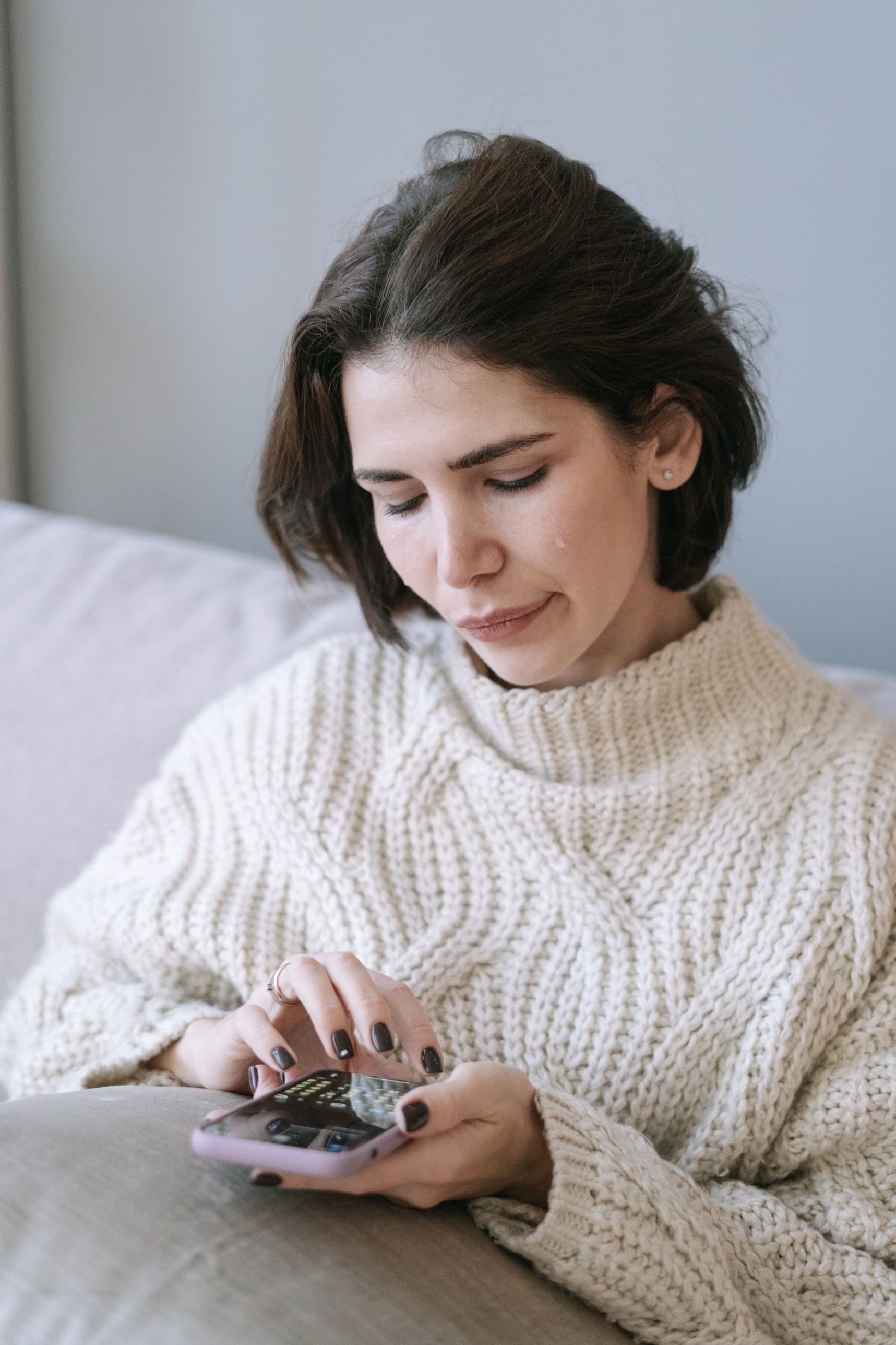 A woman on a couch in a thick sweater, typing on her phone, a tear on her cheek.