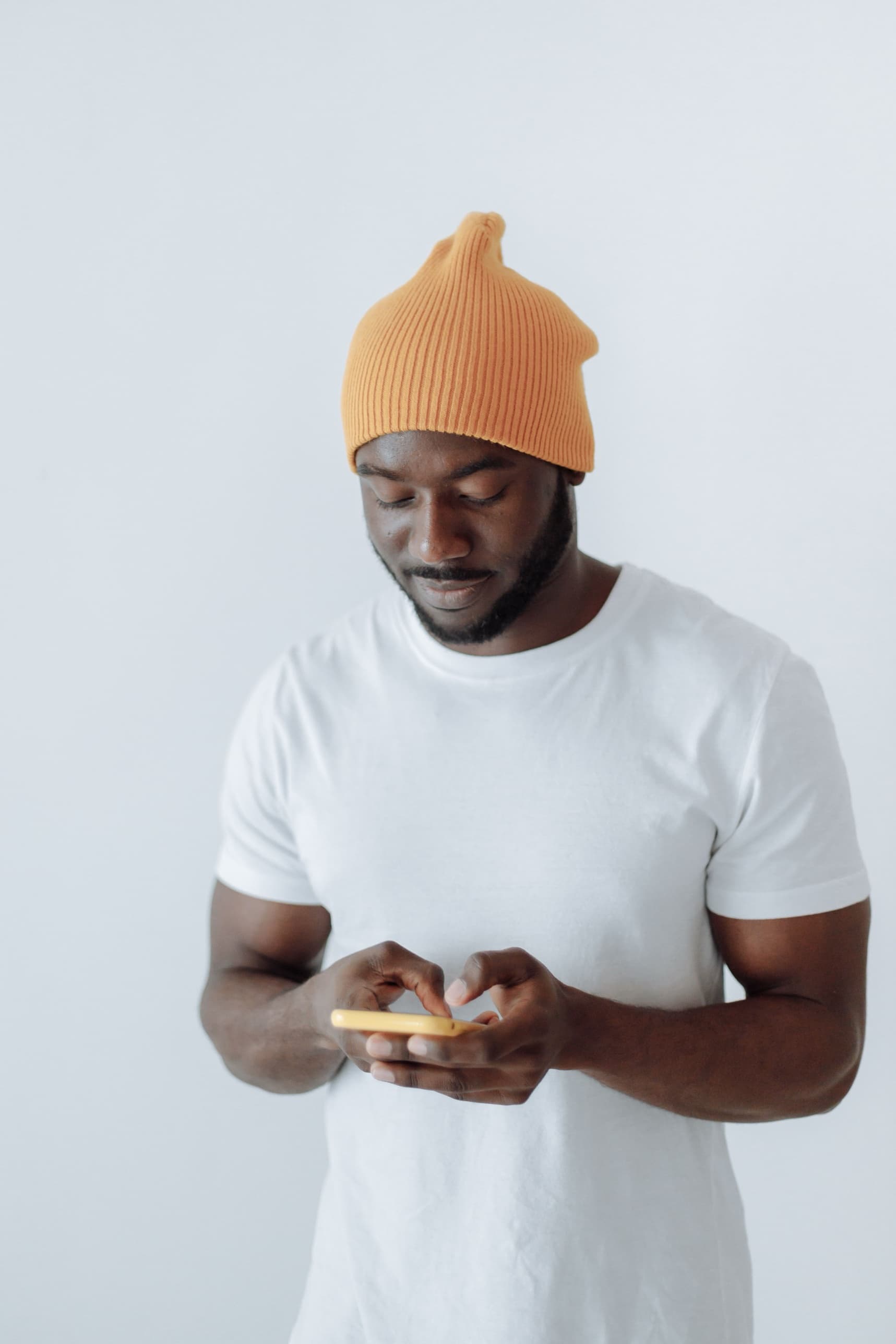 A man in an orange beanie typing on his phone against a plain wall, half smiling.
