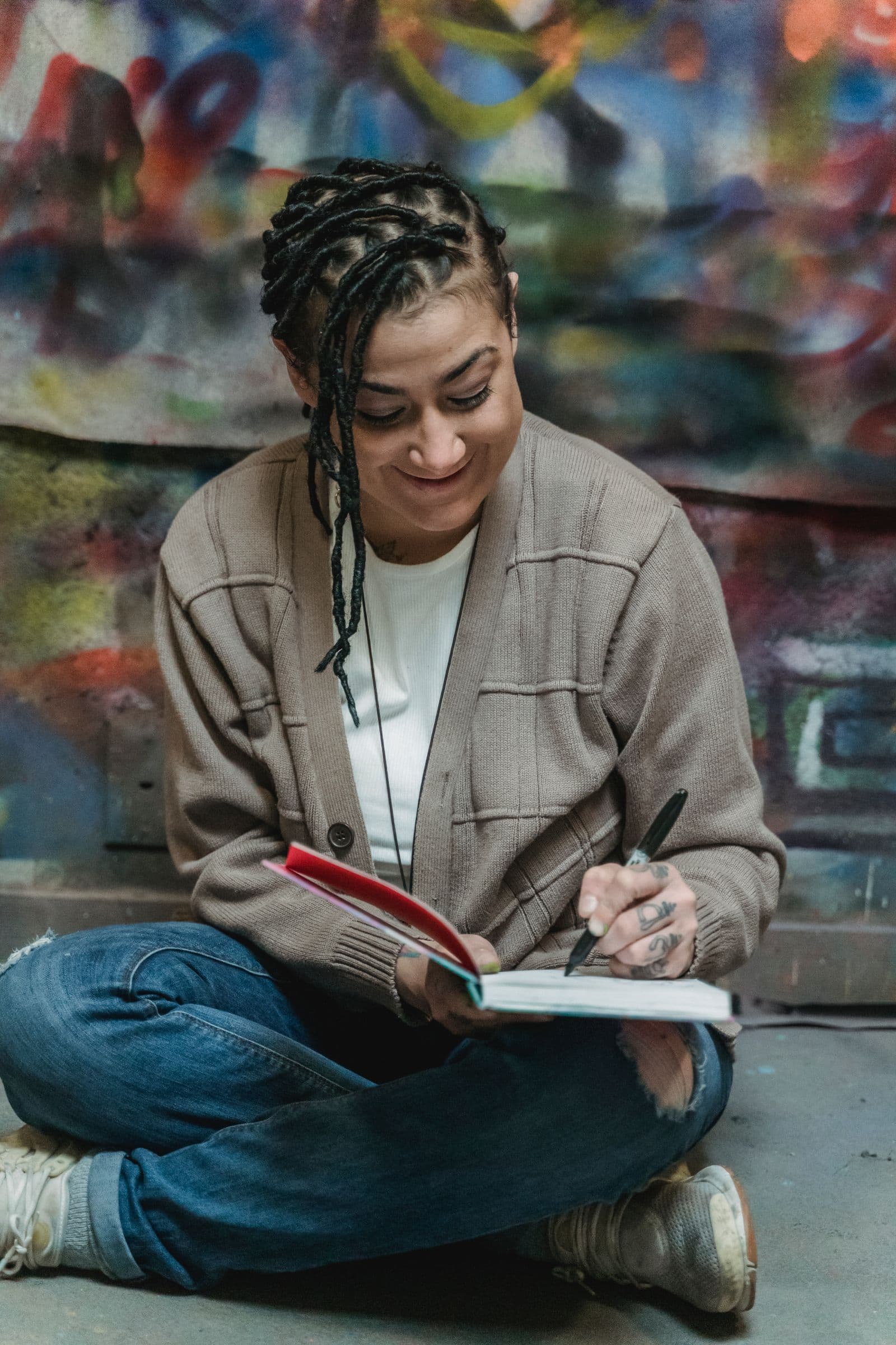 A woman sitting cross-legged on the floor against a painted wall, writing in a notebook and smiling.
