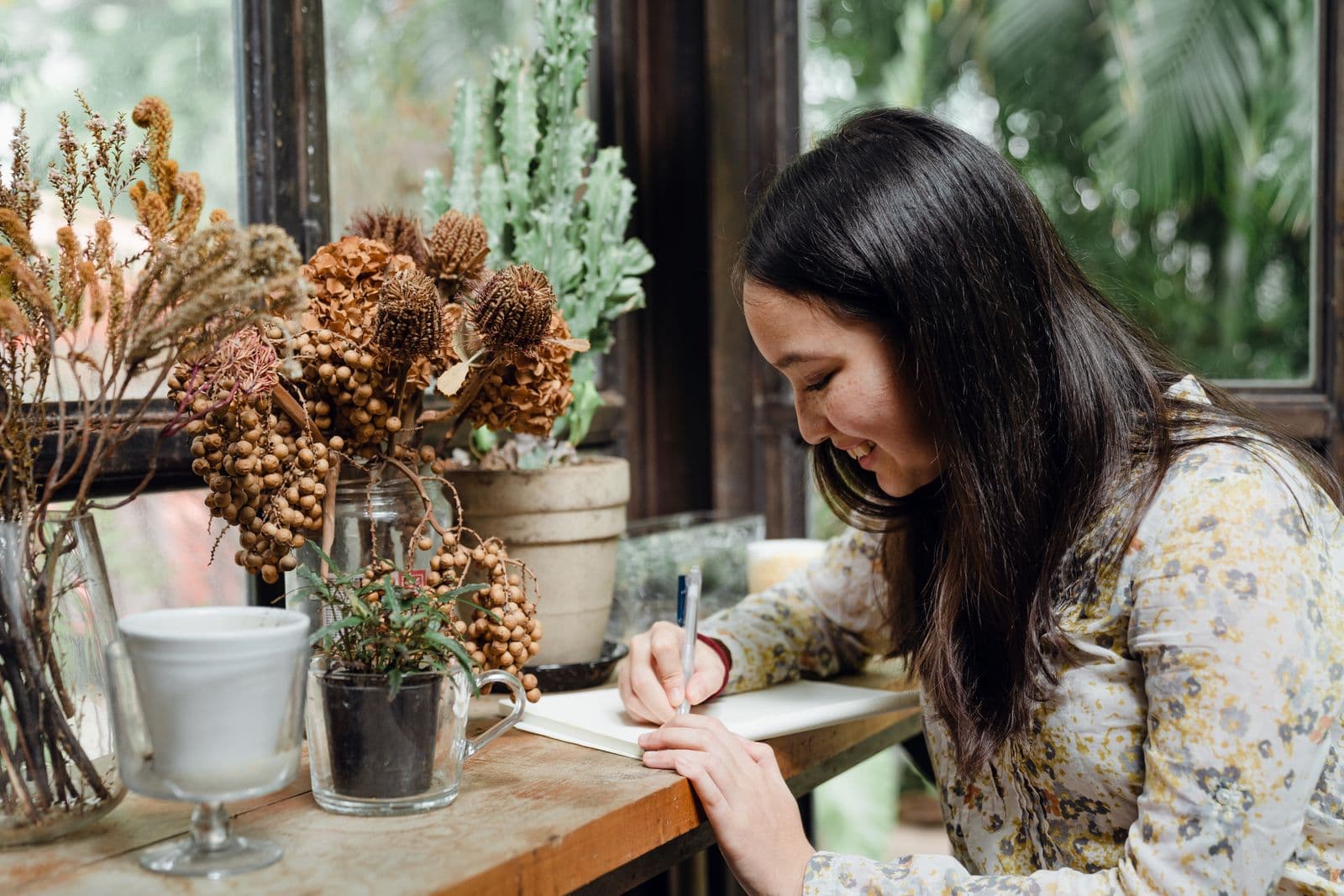 A woman writing in a notebook at a window, smiling to herself, plants on the sill beside her.